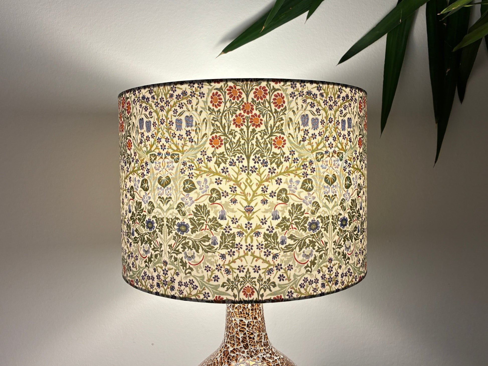 A table lamp with a drum shade featuring a dense floral and botanical pattern in cream, green, orange, and blue, set against a neutral wall with a plant leaf in the upper right.