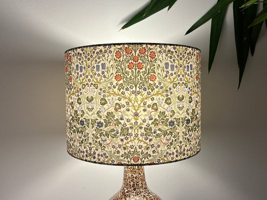 A table lamp with a drum shade featuring a dense floral and botanical pattern in cream, green, orange, and blue, set against a neutral wall with a plant leaf in the upper right.