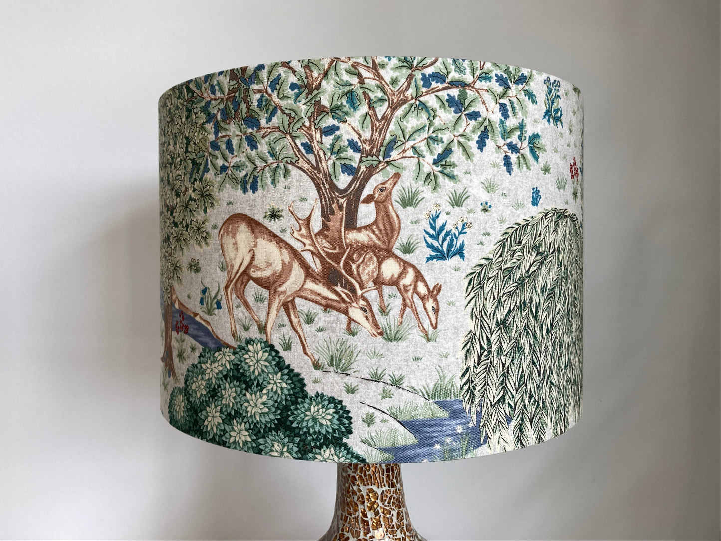 A cylindrical table lamp shade featuring a classic William Morris pattern landscape print with deer, trees, and foliage in muted blue, green, and brown tones on an off-white background, mounted on a patterned metallic base against a plain light gray wall.