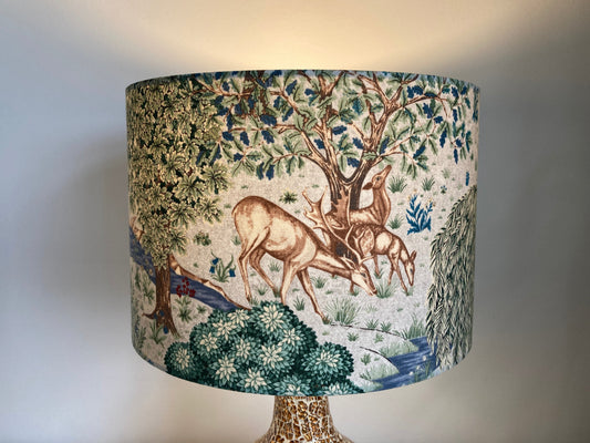 A fabric drum lampshade featuring a classic toile-style print of deer, trees, and foliage in muted blue, green, and beige tones, shown lit against a plain light gray wall. William Morris The Brook fabric in a light neutral colourway, made into a drum lampshade.